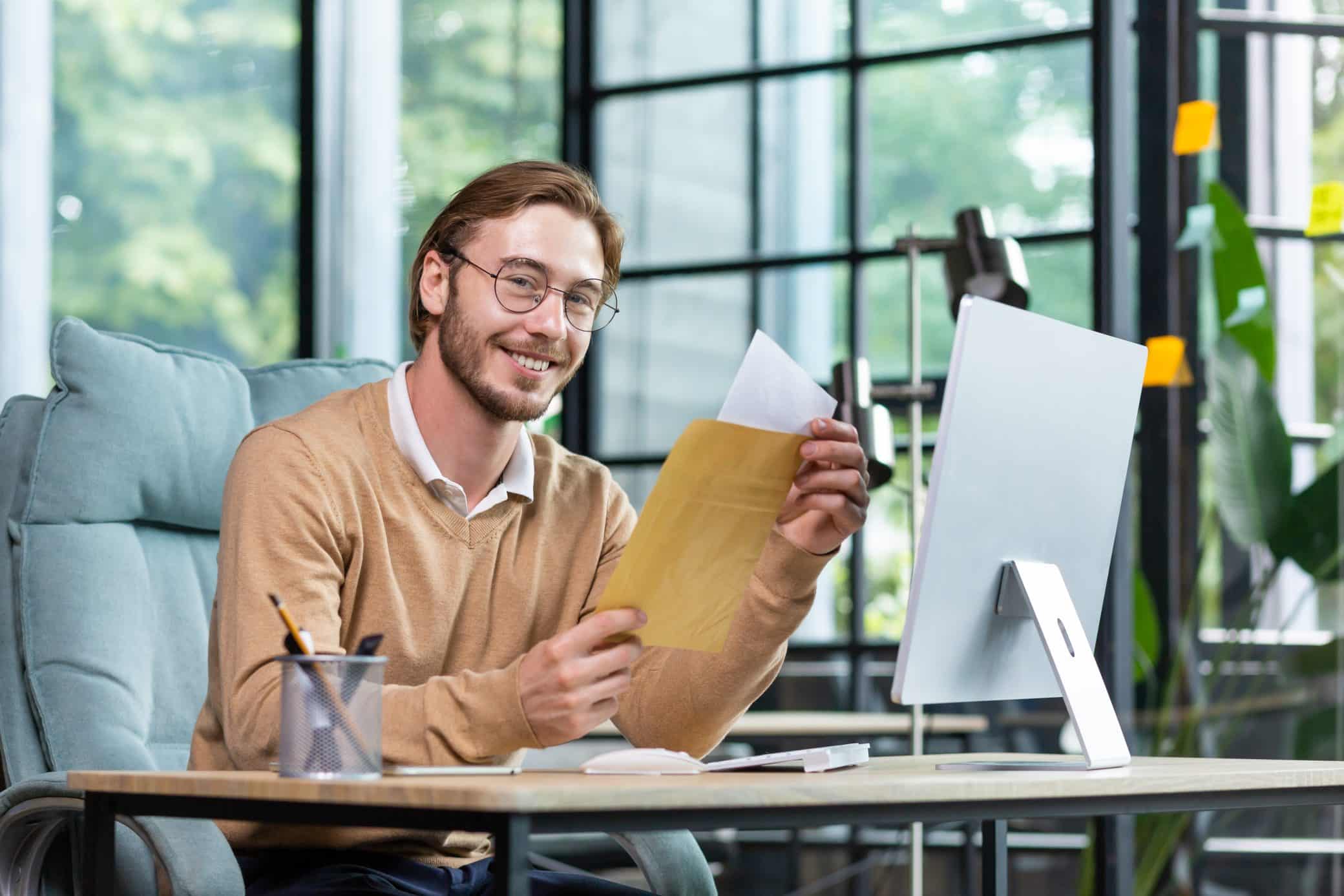 A Young Male Businessman Sits In The Office, Received And Holds An Envelope With A Letter In His Hands. Happy Smiling At Camera, Successful Deal, Financial Reward, Good News