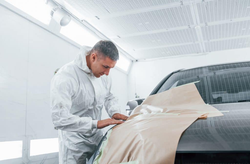 Covering Car By Brown Cloth. Caucasian Automobile Repairman In Uniform Works In Garage