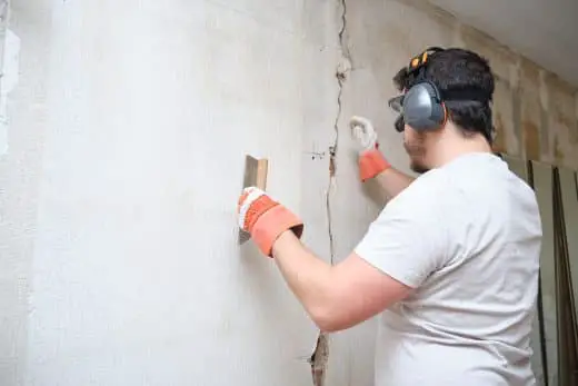 Construction Worker Repairing A Crack, Plastering Cement On Wall. Builder Wearing Safety Gloves, Glasses And Earmuffs.